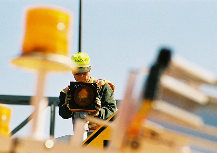 Man wearing MnDOT safety gear holds up a signal lighting component
