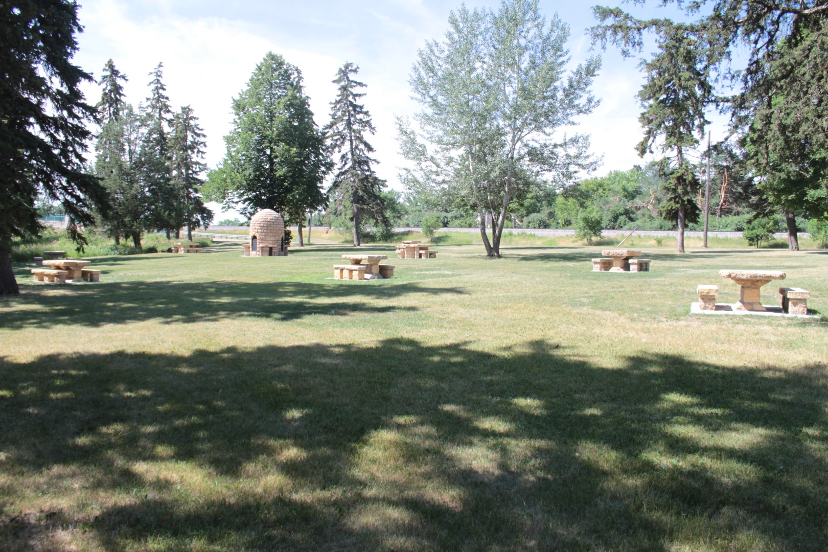 grass picnic area with six spaced out stone picnic tables, trees at edges and the “beehive” oven near the back of the view 