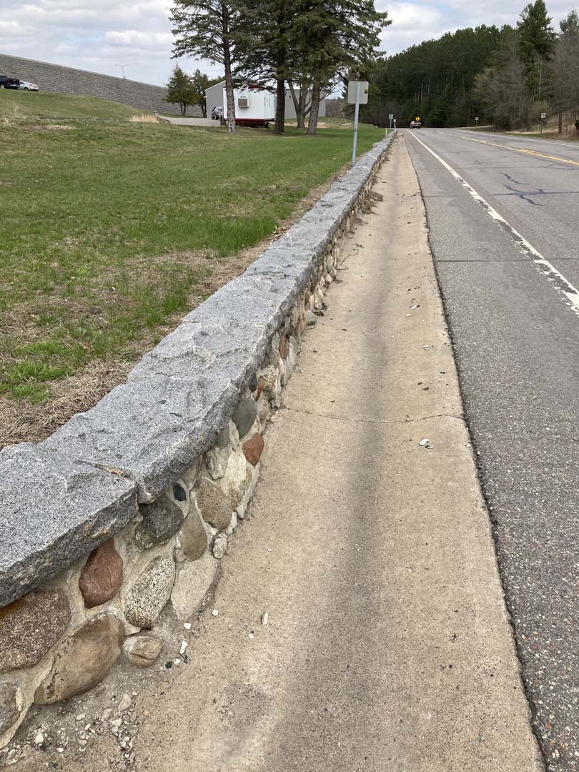 view straight down road with low retaining wall with rough grey stone cap and cobblestone face on left, concrete gutter in center and road on right. Evergreen trees in background where road features converge