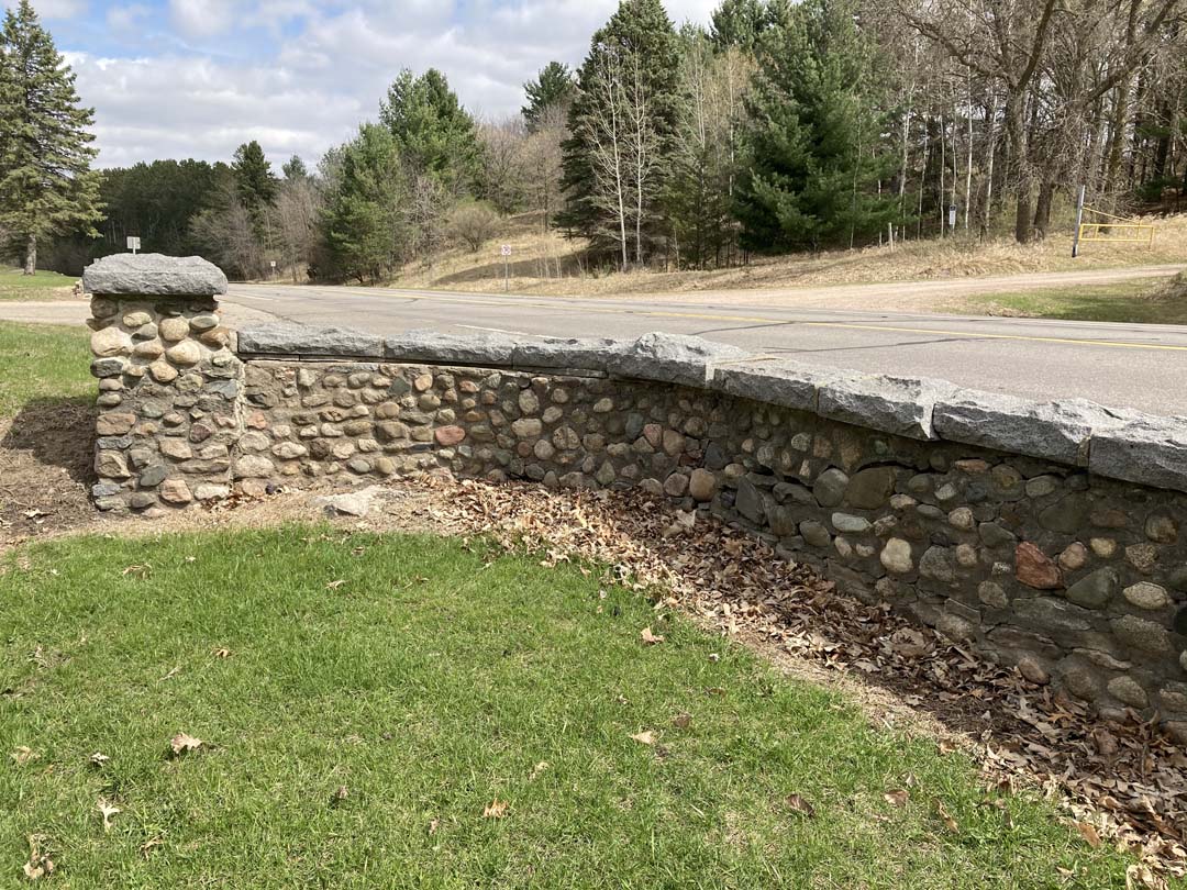 grass and retaining wall curved to taller end pier. Wall is constructed of rounded cobblestones and cut stones with rustic caps. Road is elevated behind wall and dry grass and evergreen and deciduous trees without leaves across road