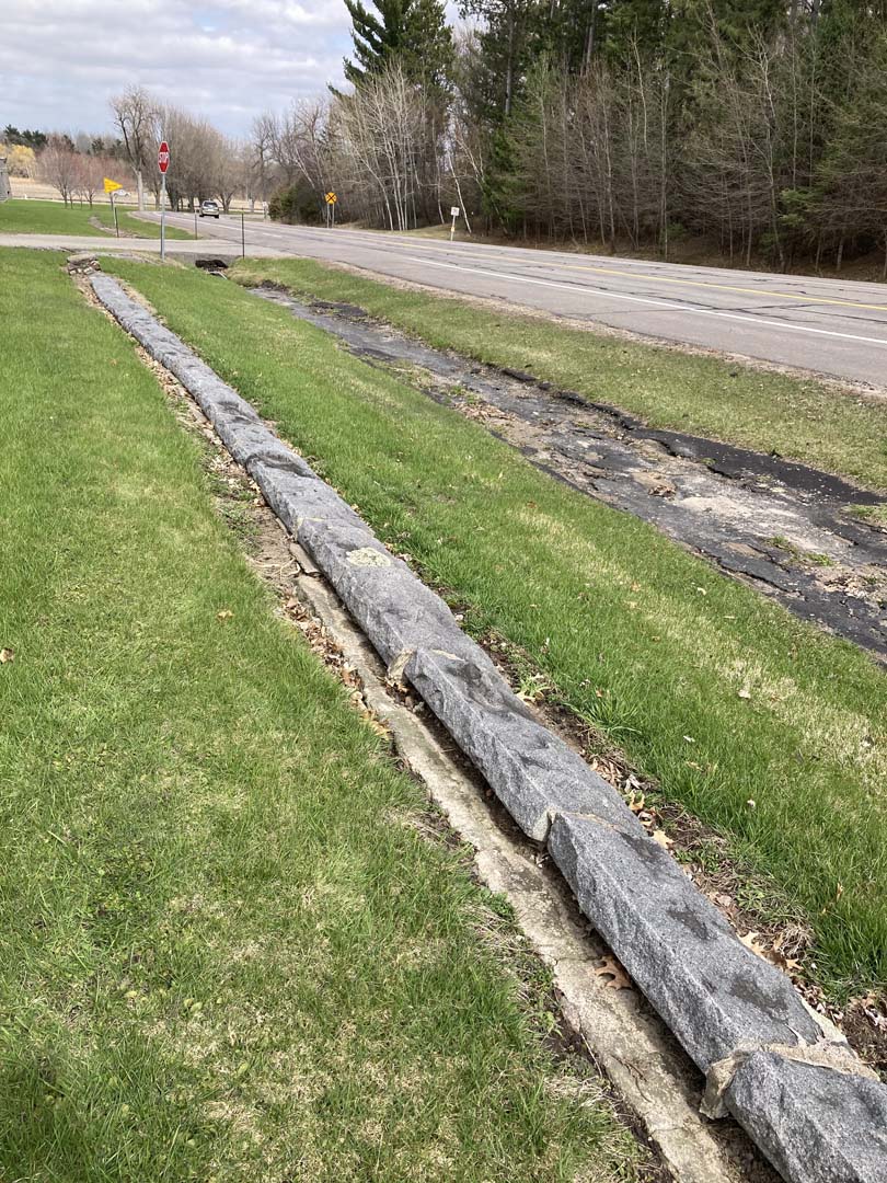low wall with only stone cap visible running through mowed green grass. Deteriorated bituminous swale runs parallel to wall a few feet away.  Road and trees visible beyond
