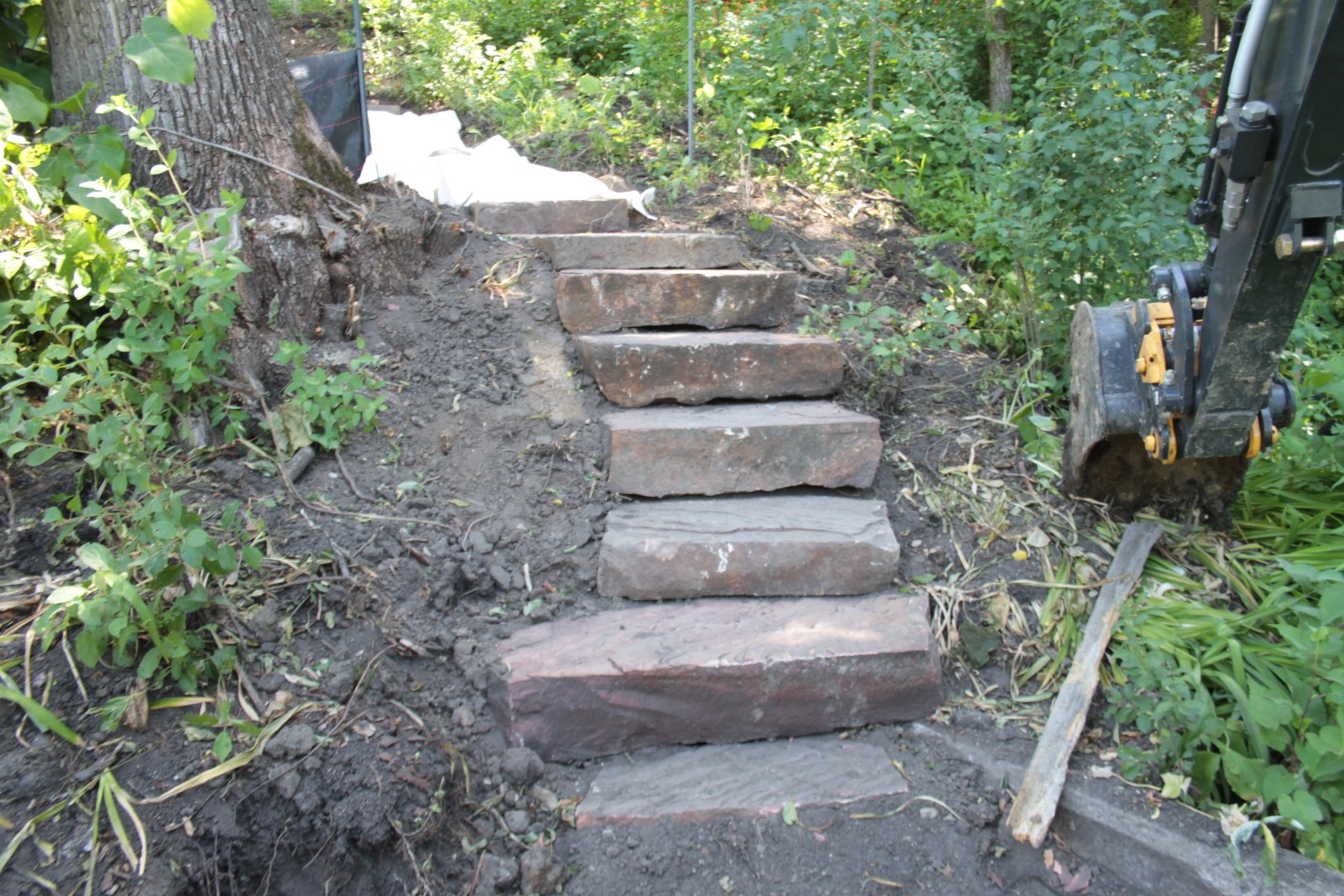 a close view of a set of stone slab steps in a wooded slope and a backhoe bucket to the right 