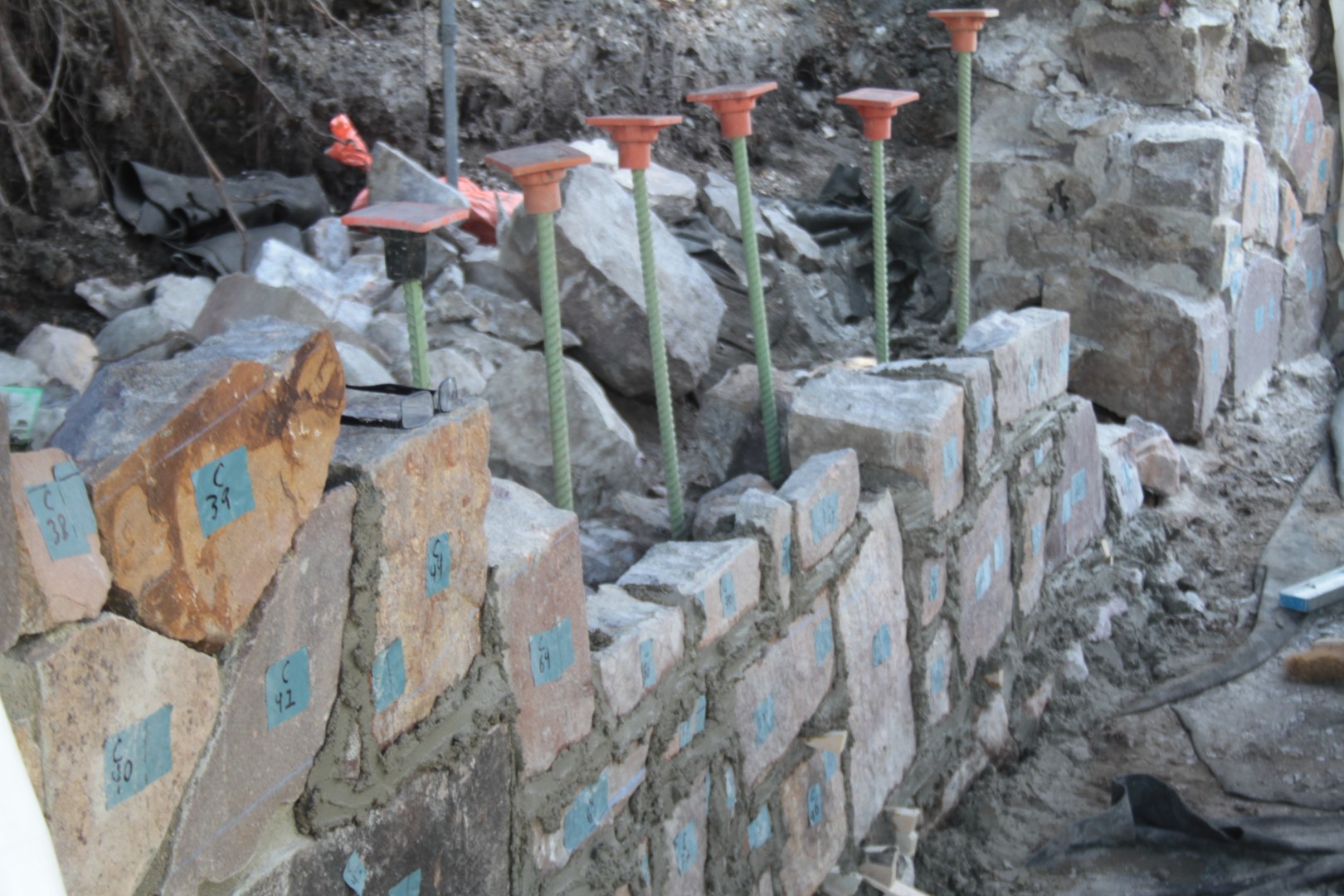 partially rebuilt stone retaining wall with wet mortar, irregular top and reinforcement rods extruding above wall with orange caps on them.