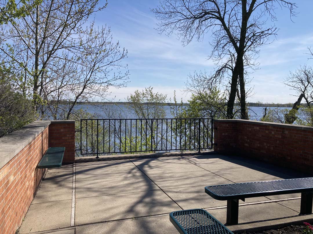 roughly square concrete surfaced plaza with benches in foreground and sides, three foot tall brick walls on sides and a metal picket railing at the far end with a view of a lake through trees with leaves in bud and blue skies with wispy clouds