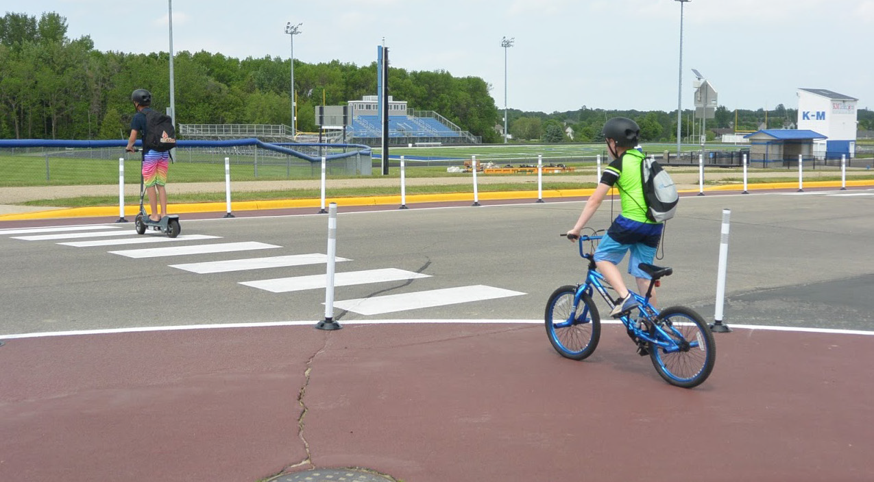 Two children bicycle and scooter through a painted curb extension with white flexible posts and a high-visibility crosswalk.