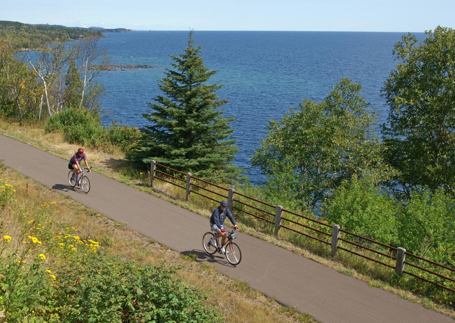 Two bicyclists ride along a paved trail overlooking Lake Superior.