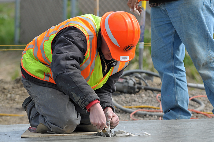 A person in yellow and orange safety gear uses a hand tool to cut into wet concrete