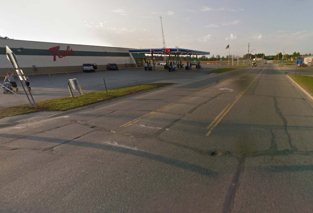 A three-lane road without sidewalks or shoulders. A person pushes a stroller through a parking lot adjacent to the right-of way.