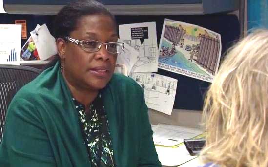 A woman in a green sweater sits at a desk as she speaks to a fellow employee.