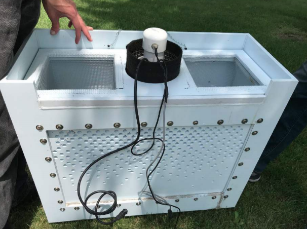 A rectangular white plastic box designed to collect street meltwater data. The box has perforated sides and a screened top. A cylindrical black-and-white instrument is mounted on the top with wire leads to transmit data. 